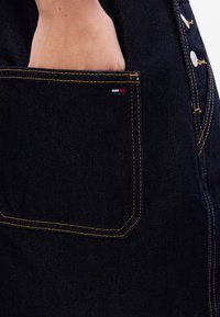 Tumši džinsi ar sānu kabatu, kurai ir dzeltenā diega apdare un neliels sarkans, balts un zils logo. Tekstūrēta auduma, pieguļoša forma.