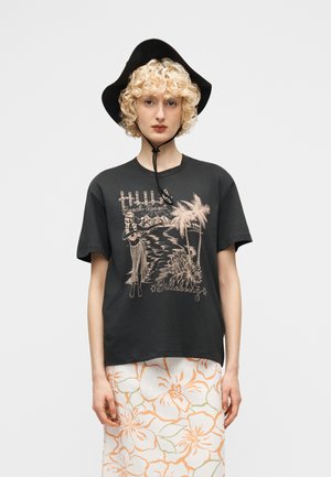 Junge Person mit lockigem blondem Haar, die einen schwarzen breitkrempigen Hut, ein schwarzes T-Shirt mit tropischem Motiv und einen weißen Rock mit orangefarbigem Blumenmuster trägt.