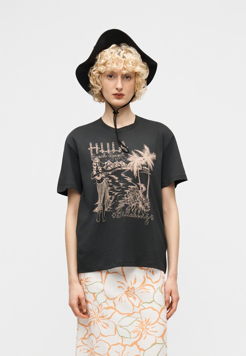 Νεαρό άτομο με σγουρά ξανθά μαλλιά που φοράει μαύρο καπέλο με πλατύ γείσο, μαύρο γραφικό μπλουζάκι με τροπικό σχέδιο και λευκή φούστα με πορτοκαλί λουλουδάτο σχέδιο.