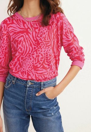 Femme portant un pull imprimé animal rose et rouge avec les manches retroussées, des colliers en or superposés et un jean taille haute bleu, une main dans la poche.