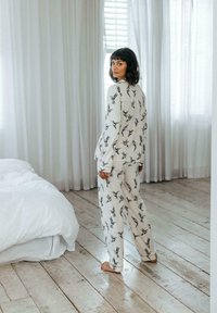 White pajama set with black bird print, featuring loose-fitting top and pants. Room has light wooden floors and sheer curtains.