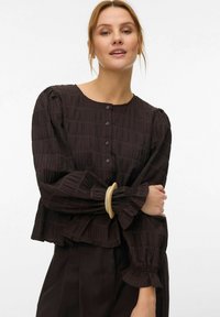 Kvinne iført en mørk, teksturert bluse med knapper og puffede lange ermer, samt et beige armbånd, stående med armene krysset og lett sminke.