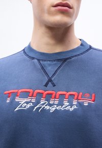 Άνδρας που φοράει μπλε φούτερ με κεντημένο λογότυπο "Tommy Los Angeles" σε κόκκινο και λευκό στο στήθος, κοντινή λήψη στον λαιμό και το κάτω μέρος του προσώπου.