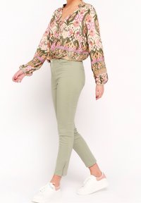 Bloes met bloemenpatroon in olijfgroen en roze, gecombineerd met olijfgroene skinnybroek met ritsen bij de enkels en witte sneakers.