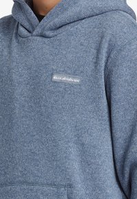Modrá fleecová mikina s texturovaným vzorem, která má přední kapsu a malé bílé logo Quiksilver na hrudi.