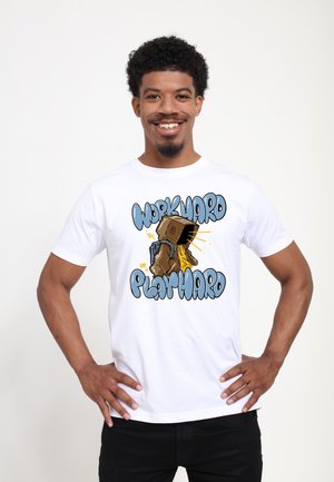 Uomo sorridente con le mani sui fianchi che indossa una maglietta bianca con una figura dei cartoni animati con testa a forma di scatola e la scritta "Work Hard Play Hard."