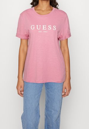 Osoba w różowej koszulce Guess z napisem "GUESS EST. 1981" oraz jasnoniebieskich dżinsach, stojąca na tle jednolitego tła.