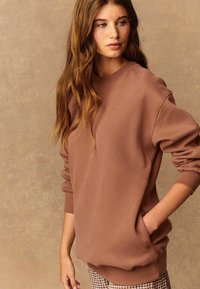 Bruine sweater met een ronde hals en geribde mouwen. De stof lijkt zacht en gestructureerd. Zak aan de zijkant. Gekoeld met geruite broek.