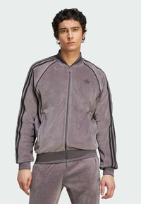 Joven con cabello corto y rizado, vistiendo un chándal de terciopelo gris de Adidas con franjas negras en las mangas y pantalones, de pie frente a un fondo liso.