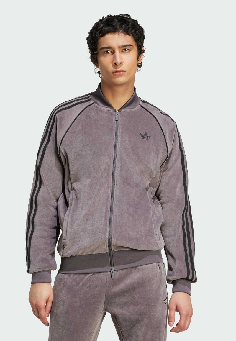 Joven con cabello corto y rizado, vistiendo un chándal de terciopelo gris de Adidas con franjas negras en las mangas y pantalones, de pie frente a un fondo liso.