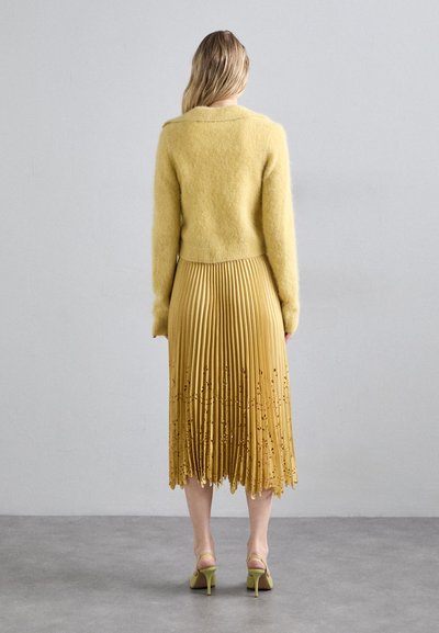 Pulover galben din pluș asociat cu o fustă plisată muștar, având detalii florale decupate și pantofi cu toc înalt galbeni.