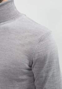 Lys grå høyhalset genser laget av strikket stoff, med en tettsittende design og subtile ribbestrikkede detaljer langs sømmene.