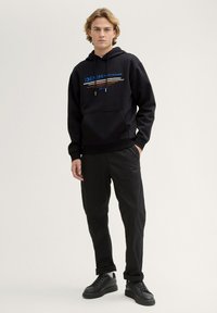 Sudadera negra con bolsillo frontal, impresión de logo colorido, capucha con cordones; combinada con pantalones negros, ligeramente ajustados y zapatillas negras.