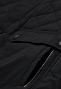 Nahaufnahme einer schwarzen gesteppte Jackentasche mit einem Klappdeckel, der mit einem Druckknopf gesichert ist, und sichtbaren Nähdetails.