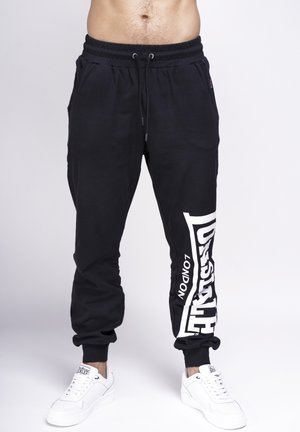 Black sweatpants with an elastic waistband and drawstring. Features a large white logo on the left leg. Paired with white sneakers.