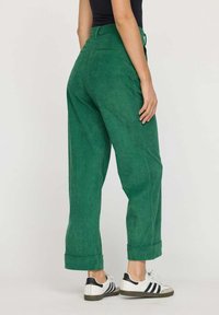 Pantalons en velours côtelé vert avec une taille haute, des poignets roulés et des poches arrière. Associés à des chaussures de sport blanches et noires.