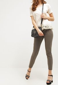 Kvinna i en vit fransad topp, olivfärgade slim fit-byxor, svarta blockklack-sandaler och bär en svart crossbody-väska.