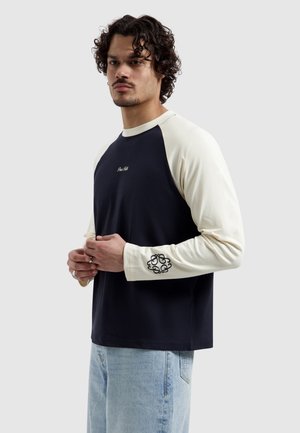 Mann mit lockigem Haar, der ein marineblaues und cremefarbenes Langarmshirt mit schwarzem Emblem am Ärmel und hellblaue Jeans trägt, steht mit verschränkten Händen.