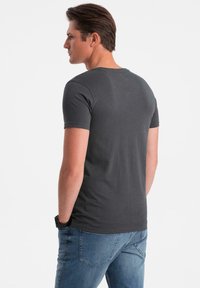 Uomo con capelli corti castani che indossa una t-shirt a maniche corte grigia scura e jeans blu, in piedi con le mani nelle tasche, rivolto a sinistra.