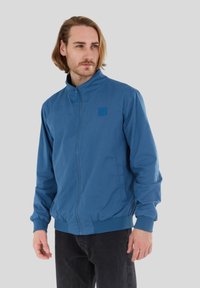 Blaues Zip-Jacke mit Stehkragen, elastischen Bündchen und minimalistischem Logo auf der Brust. Hergestellt aus glattem, leichtem Stoff.