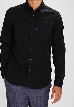 Mężczyzna ubrany w czarną, zapinaną na guziki koszulę z długim rękawem z kieszeniami z klapkami oraz teksturowane szare spodnie, stojący z jedną ręką przy boku.