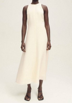 Maxi-jurk in een zachte, off-white stof, met een hoge halslijn, mouwloos ontwerp en een ruwe zoom. Gedragen met zwarte sandalen.