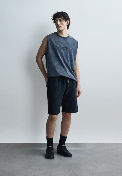 Νέος άνδρας φοράει αμάνικο ξεθωριασμένο γκρι πουκάμισο "Off-White", μαύρο σορτς, μαύρες κάλτσες και μαύρα αθλητικά παπούτσια, στέκεται μπροστά σε έναν απλό τοίχο.