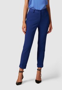 Femme portant un pantalon ajusté bleu roi longueur cheville avec des ourlets retroussés et des escarpins noirs à bout pointu avec des brides à la cheville.