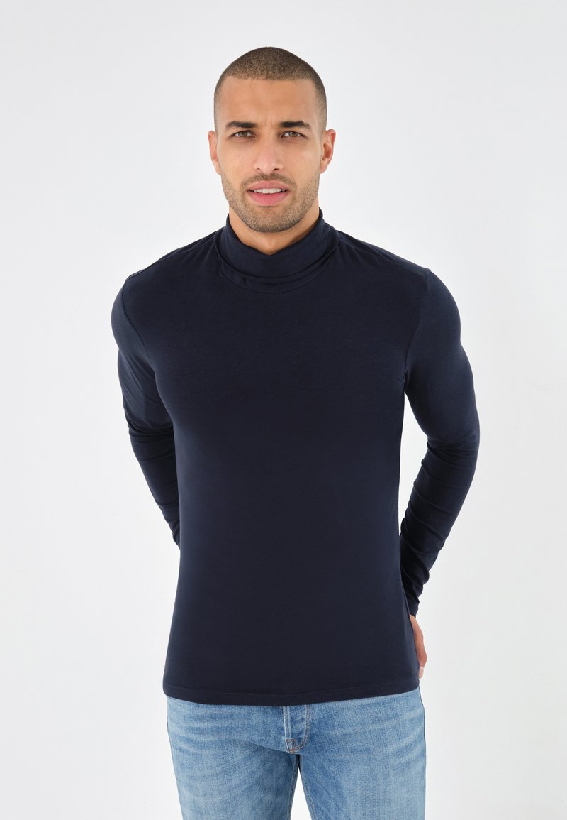 Cămașă din bumbac cu mânecă lungă și guler înalt, de culoare bleumarin, realizată dintr-un material neted și elastic. Prezintă un design ajustat, purtată cu blugi de culoare albastru deschis.