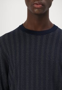 Maglione blu navy con un motivo a spina di pesce testurizzato e fili metallici. Collo a costine e tessuto morbido.