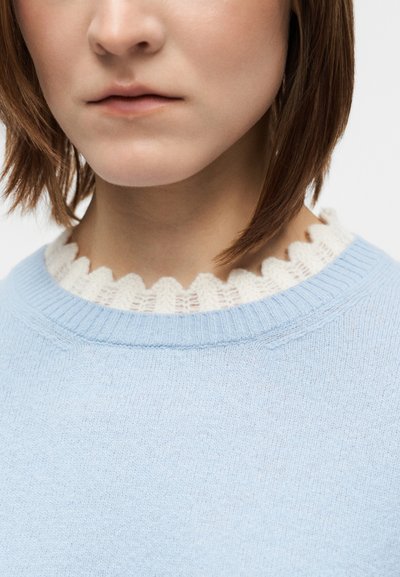 Hellblauer Pullover mit einem gerippten Rundhalsausschnitt und einem kontrastierenden weißen, gewellten Spitzenbesatz. Weiche Textur, figurbetontes Design, minimalistische Details.