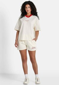 Camiseta de algodón de color crema, oversized, con un cuello en V rojo, combinada con unos pantalones cortos de crema a juego que presentan un texto bordado en el lado izquierdo.