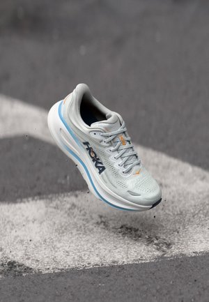 Chaussure de course Hoka gris clair avec des accents bleus et orange flottant au-dessus d’un bitume marqué d’une ligne blanche.