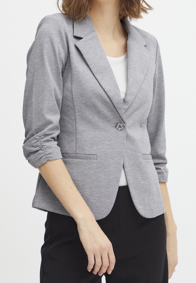 Grå, lätt kavaj med slagkrage, enkel knappknäppning och rynkade trekvartsärmar. Har två framfickor.