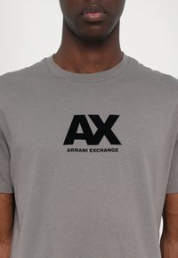 Pilkos spalvos medvilninė marškinė su juodu logotipu "AX ARMANI EXCHANGE" krūtinėje. Trumpi rankovių ir apvalus iškirpimas.