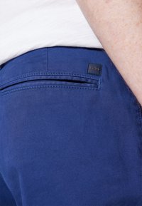 Mörkblå byxor i slätt tyg, med en bakficka som har en liten logotag i svart läder. Sömmar är synliga.