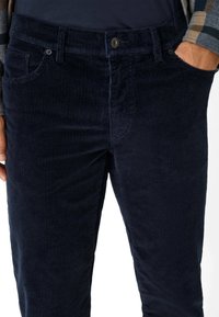 Donkerblauwe corduroypantalons met een geribbelde textuur, vijf zakken en een sluiting met een knoop aan de voorkant. Naden en stiksels zijn zichtbaar.