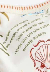 Text na tkanine s poetickými riadkami o slnku a nebi, s ilustráciou červenej morskej mušle a pleteným okrajom v krémovej a oranžovej farbe.