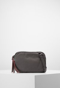 Šedá kožená crossbody taška se zipovým uzavřením, s hladkým povrchem a červenými dekorativními třásněmi po stranách. Kompaktní obdélníkový tvar.