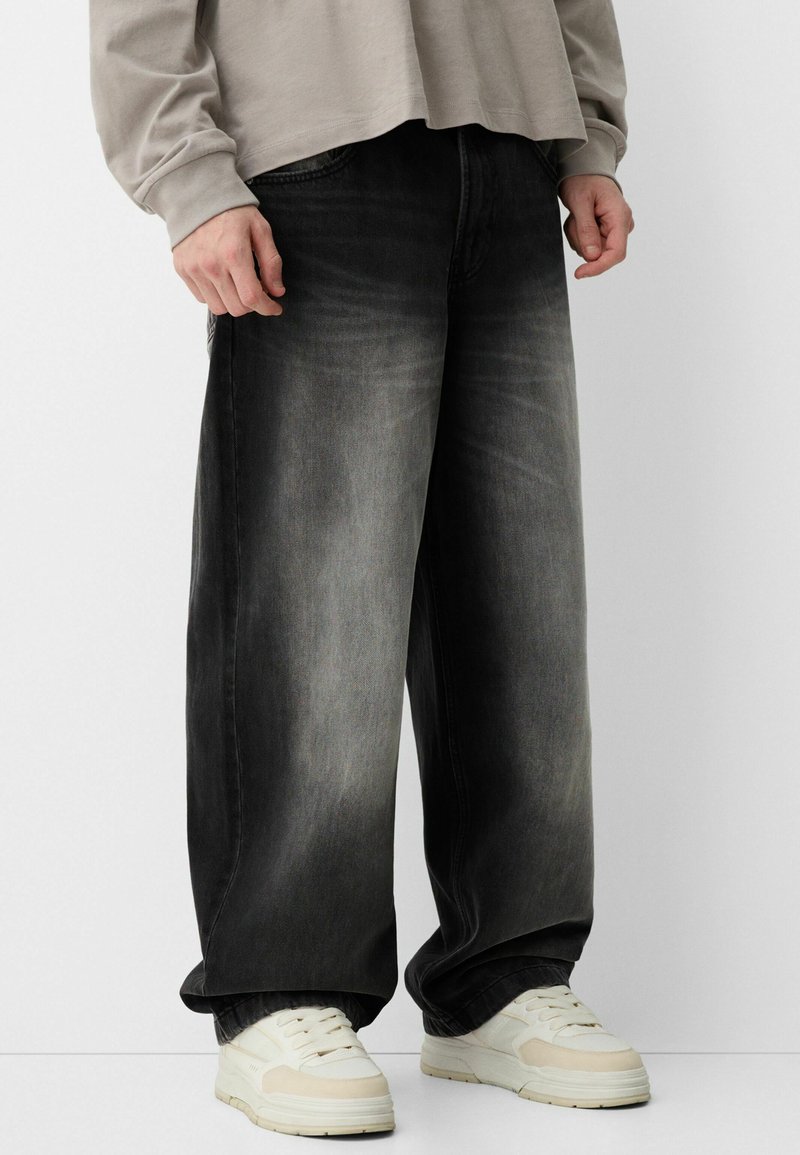 Pohodlné černé džíny z denimu s vybledlým efektem, typický pětikošilový design, spárovány s bílými teniskami. Tkanina vykazuje jemné variace textury.