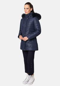 Chaqueta acolchada azul marina con capucha de piel sintética, cierre de cremallera y detalles de botones. Brillo texturizado, diseño a la altura de la cadera, combinada con pantalones negros ajustados y zapatillas blancas.