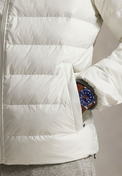 Biela prešívaná bunda s horizontálnym prešívaním, vybavená zipsom a predným vreckom. Modré hodinky vyčnievajú z vnútorného vrecka na sivom materiáli.