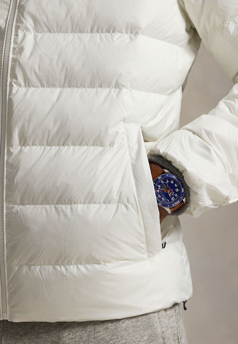 Doudoune blanche avec couture horizontale, dotée d'une fermeture éclair et d'une poche avant. Une montre bleue dépasse d'une poche intérieure sur un tissu gris.