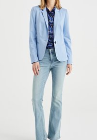 Ljusblå blazer med slagkrage, en svart knapp och två framfickor, parat med en mönstrad skjorta och ljusblå flare-jeans.