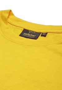 Gelbes Baumwoll-T-Shirt mit Rundhalsausschnitt. Verfügt über ein kleines Etikett mit dem Wort "Switcher" und eine Nahtdetail um den Kragen.