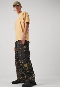 Oransje t-skjorte med korte ermer, cargo-bukser i svart og kamuflasjemønster, svarte joggesko, olivengrønn caps, stående posisjon mot et nøytralt bakgrunn.