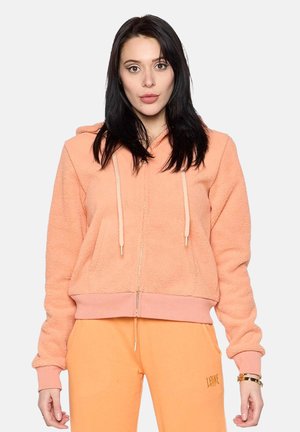 Jeune femme aux longs cheveux foncés portant un sweat à capuche pêche zippé et un pantalon assorti avec un petit logo sur la cuisse gauche.