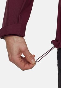 Camicia a maniche lunghe color mogano con tessuto strutturato; mano che afferra un cordino regolabile sull'orlo per il restringimento; polsino con dettaglio a linguetta visibile.