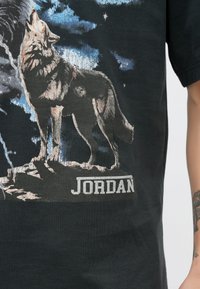 Juoda marškinėliai su kaukiančio vilko piešiniu, detaliai atliktu pilka ir balta spalvomis. Apačioje paryškintu šriftu užrašytas žodis „JORDAN“.