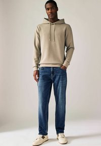 Lichtbruin hoodie met een trekkoord, gecombineerd met blauwe spijkerbroeken en crèmekleurige sneakers. Gladde textuur met een ontspannen pasvorm.